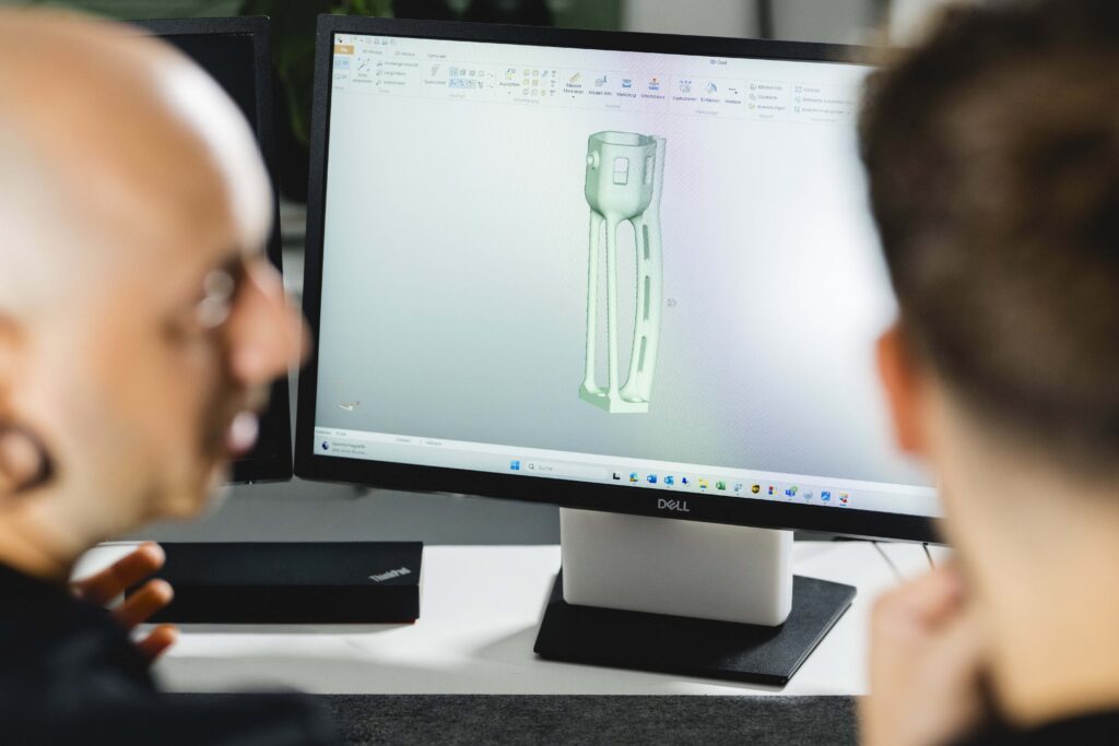 Zwei Mitarbeiter sitzen vor einem Computerbildschirm auf dem die CAD-Konstruktion eines SLS 3D-Druckbauteils zu sehen ist.