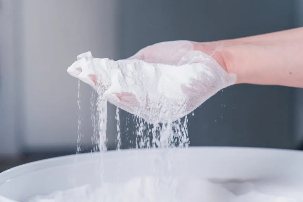 A hand holds white SLS 3D printing powder over a bucket.