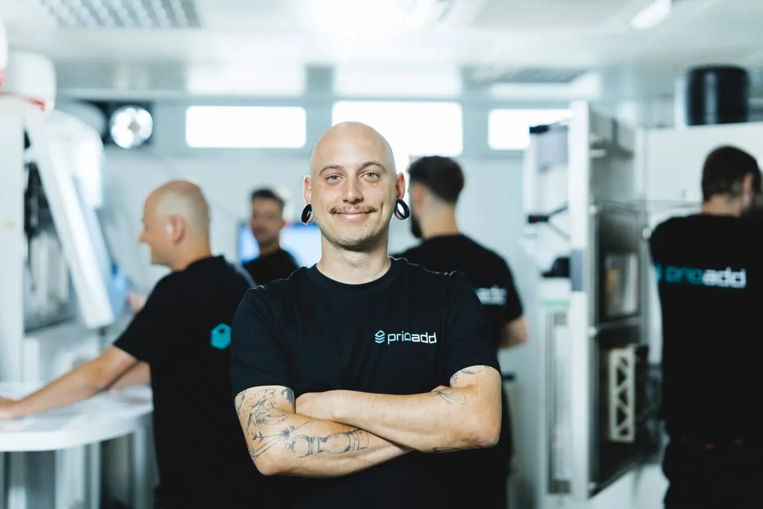 L’équipe de prioadd travaille sur des machines d’impression 3D SLS. Un collaborateur se tient au premier plan, les bras croisés.